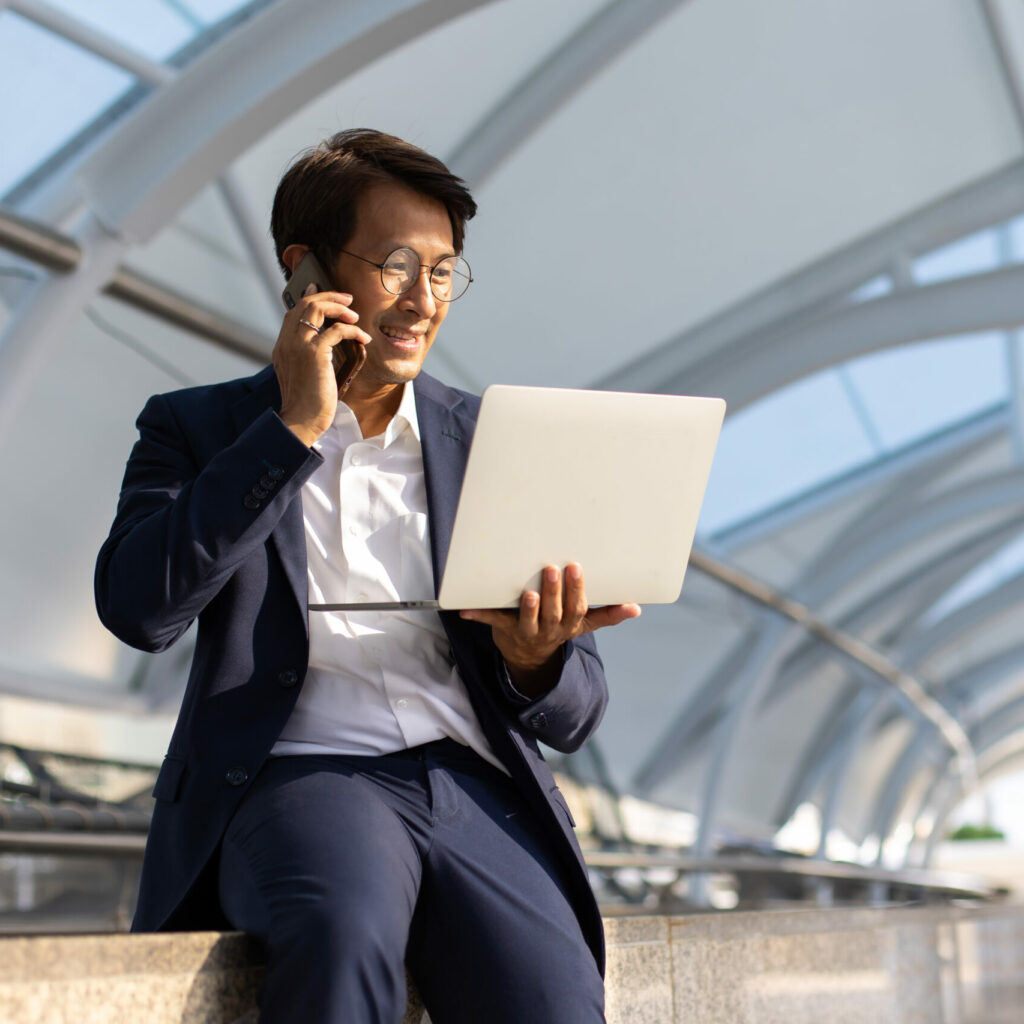 Asian confidently business man in glasses and suit working on laptop and mobole phone outdoor in city.